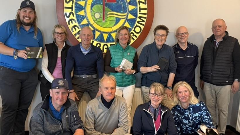Gruppenfoto in einem Clubraum mit zehn Personen und zwei Hunden, um ein Logo des Ostsee Golfclubs gebannt.