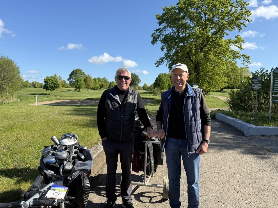 Zwei M&auml;nner stehen auf einem Golfplatz, neben ihnen eine Golftasche mit Schl&auml;gern, umgeben von gr&uuml;ner Wiese und B&auml;umen.