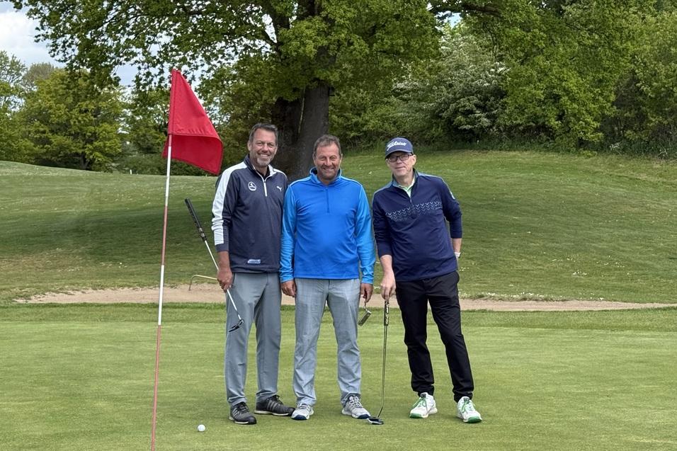 Drei Golfer posieren auf dem Gr&uuml;n neben einer roten Fahne, umgeben von B&auml;umen und einer gepflegten Landschaft.
