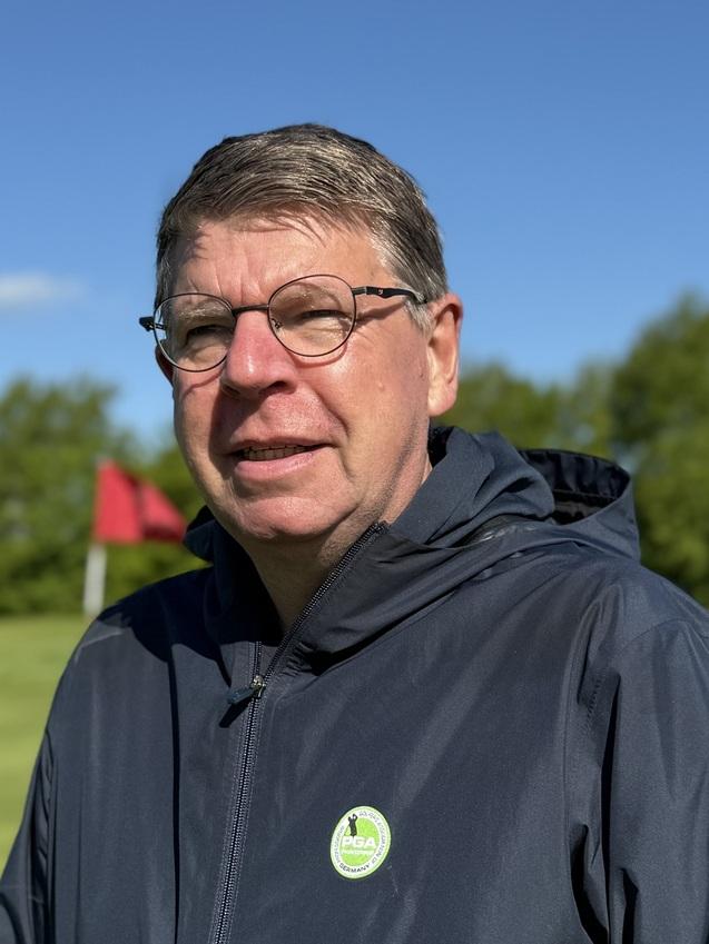 Mann in einer dunklen Jacke steht auf einem Golfplatz mit rot-weißem Fahnenmast im Hintergrund und blauem Himmel.
