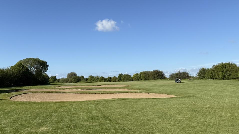 Gr&uuml;nes Feld mit zwei Sandfl&auml;chen und einem Golfwagen im Hintergrund unter klarem, blauem Himmel.