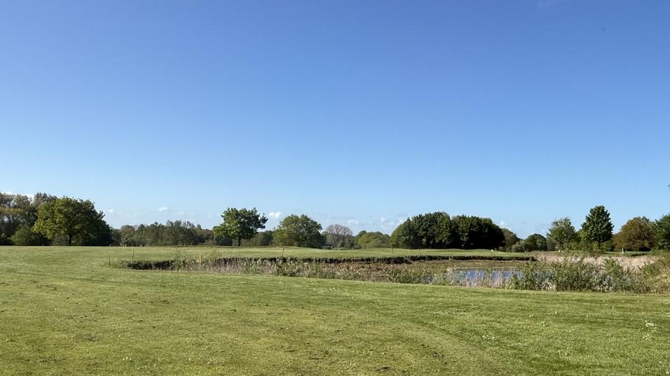 Weite Landschaft mit gr&uuml;nem Gras, B&auml;umen und einem Wasserlauf unter blauem Himmel mit ein paar Wolken.