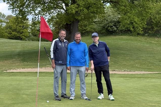 Drei Golfer posieren auf dem Grün neben einer roten Fahne, umgeben von Bäumen und einer gepflegten Landschaft.