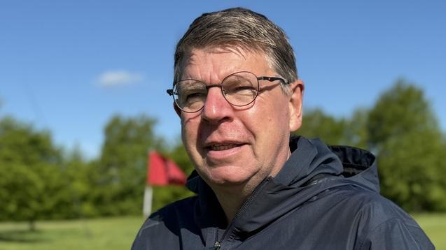 Mann in einer dunklen Jacke steht auf einem Golfplatz mit rot-wei&szlig;em Fahnenmast im Hintergrund und blauem Himmel.