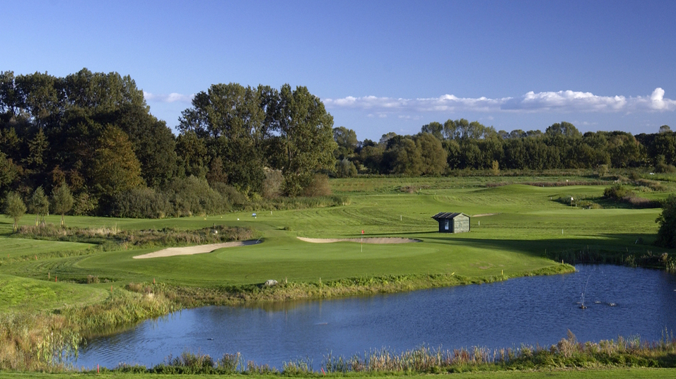 Panoramablick auf einen Golfplatz mit Teich, B&auml;ume und klarem Himmel im Hintergrund.