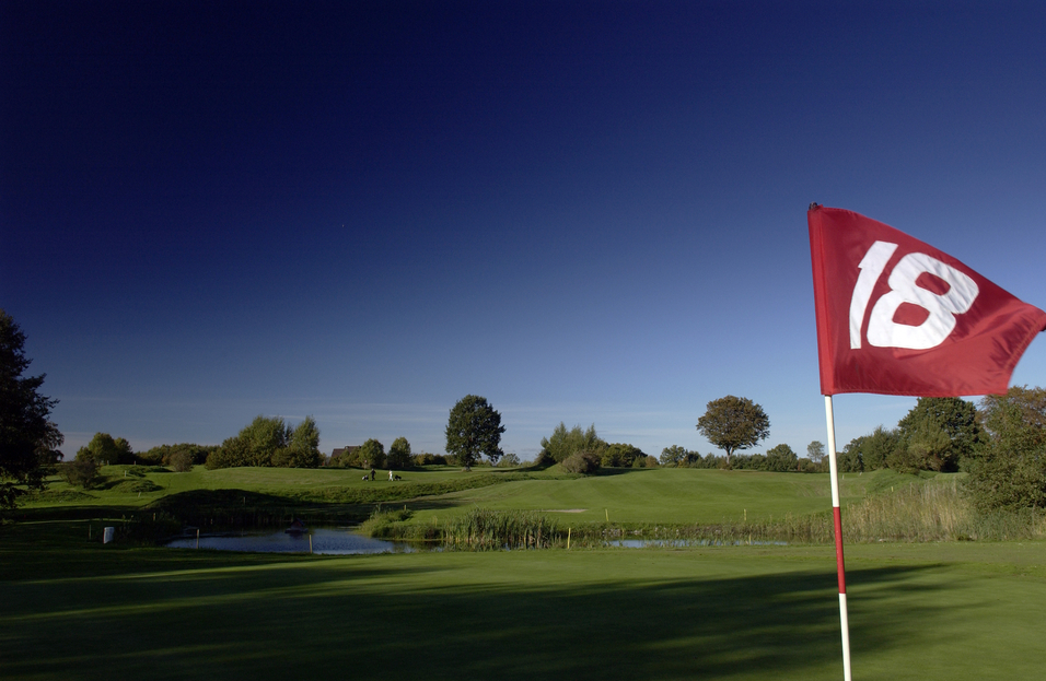 Ansicht eines Golfplatzes mit einer Fahne am 18. Loch, B&auml;ume und Wasser im Hintergrund unter blauem Himmel.