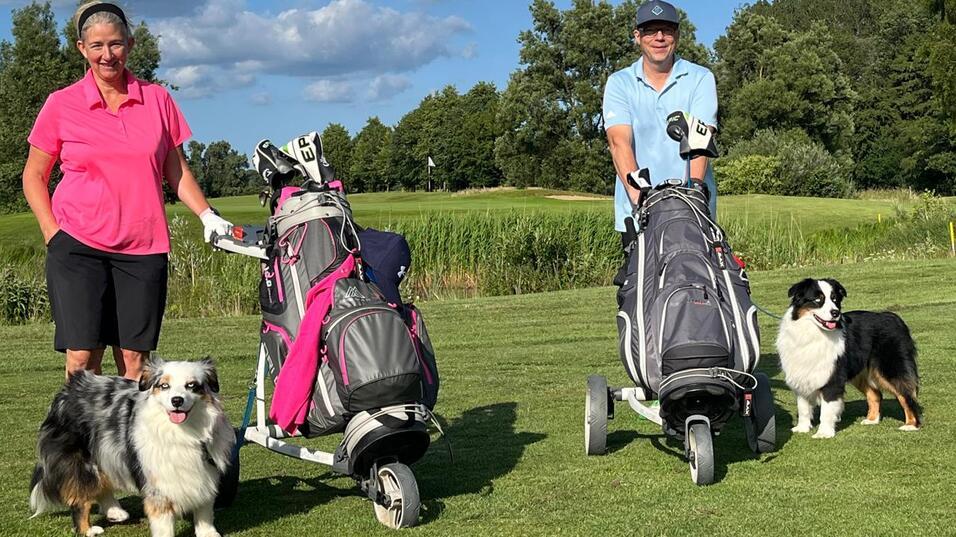 Zwei Golferinnen mit Golftaschen stehen auf einem Platz, begleitet von zwei Hunden, unter einem blauen Himmel mit Wolken.