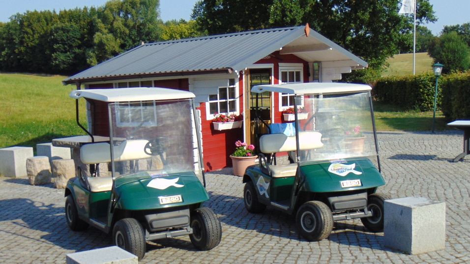 Zwei Golfcarts parken auf einem gepflasterten Platz vor einem kleinen roten Geb&auml;ude und einer gr&uuml;nen Wiese.