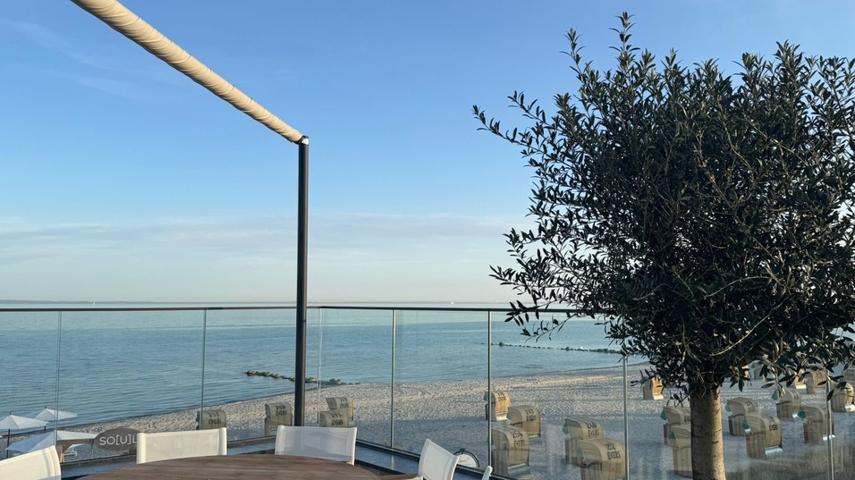 Terrasse mit Rundtisch und Olivenbaum, Blick auf das Meer und Liegen am Strand im Hintergrund.