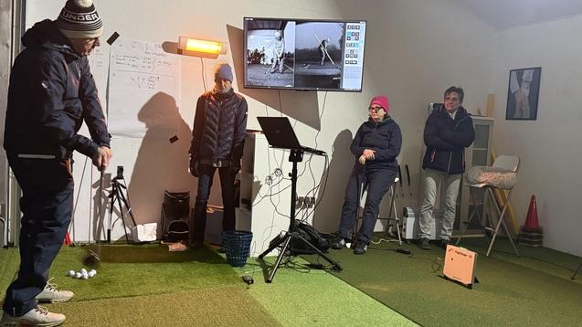 Person beim Golftraining in einer Indoor-Anlage, Zuschauer beobachten im Hintergrund. Bildschirm zeigt Trainingsanalysen.