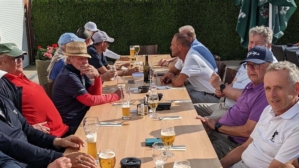 Gruppe von M&auml;nnern sitzt an einem Tisch drau&szlig;en, Biergl&auml;ser und Speisen sind auf der Tischfl&auml;che verteilt.