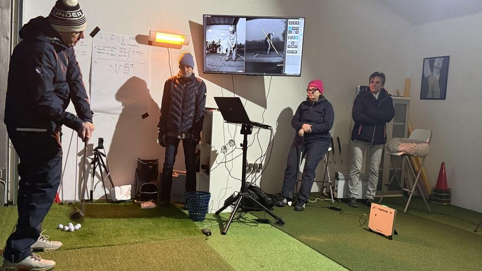 Person beim Golftraining in einer Indoor-Anlage, Zuschauer beobachten im Hintergrund. Bildschirm zeigt Trainingsanalysen.