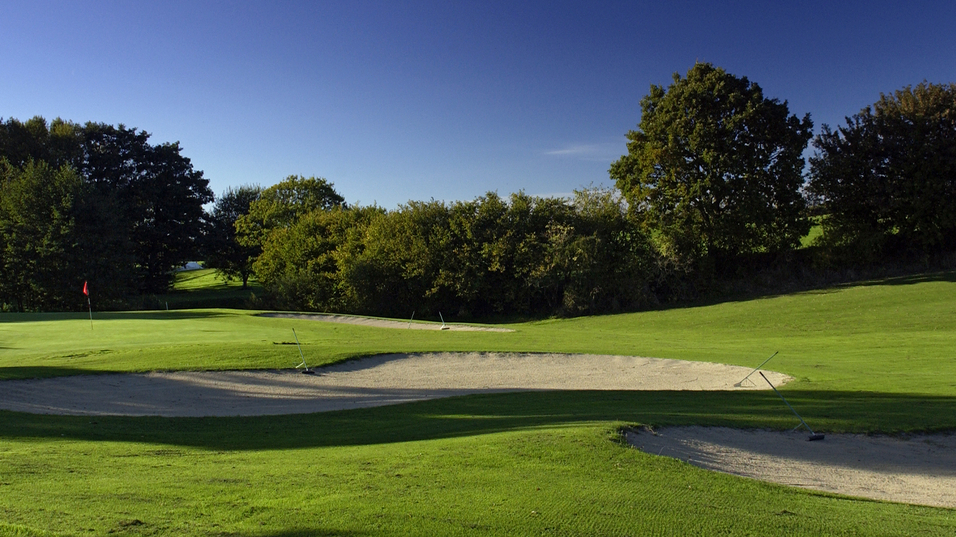 Golfplatz mit Sandbunker, gepflegtem Rasen und B&auml;umen unter klarem, blauem Himmel.