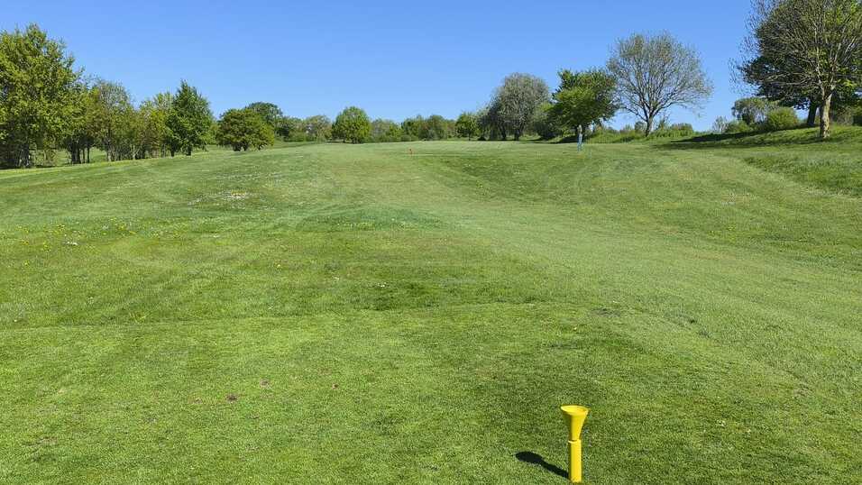 Blick auf einen Golfplatz mit gr&uuml;nem Rasen und gelbem Abschlagtee vor sanften H&uuml;geln und B&auml;umen im Hintergrund.