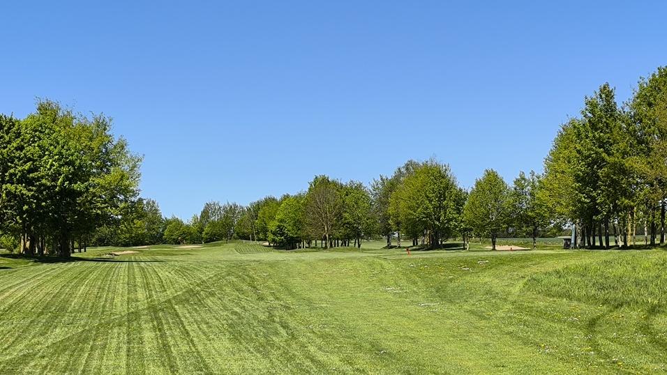 Blick auf eine gepflegte Golfanlage mit gr&uuml;nem Rasen und B&auml;umen unter blauem Himmel.