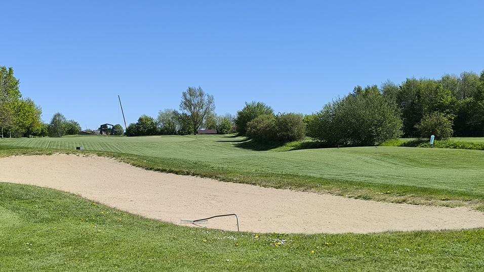 Blick auf eine Golfbahnanlage mit einem Bunker und saftigem Gr&uuml;n unter blauem Himmel.