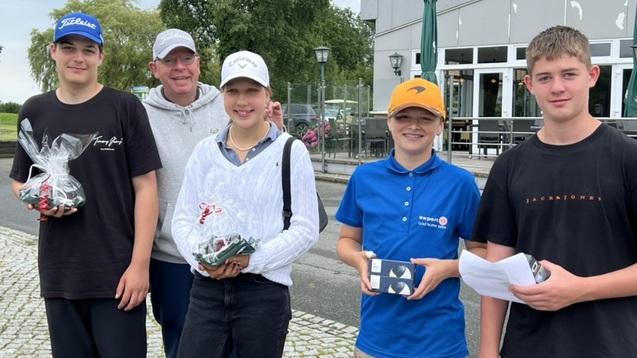 Gruppe von vier Jugendlichen und einem Erwachsenen, die Pokale nach einem Wettbewerb l&auml;cheln, vor einem Caf&eacute; im Freien.