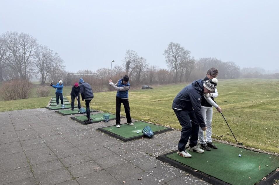Sechs Golfer &uuml;ben auf separate Abschlagb&ouml;den auf einem nebligen Golfplatz.