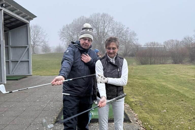 Zwei Personen stehen auf einer Golf Driving Range, halten Golfschl&auml;ger und posieren l&auml;chelnd vor einer bew&ouml;lkten Kulisse.