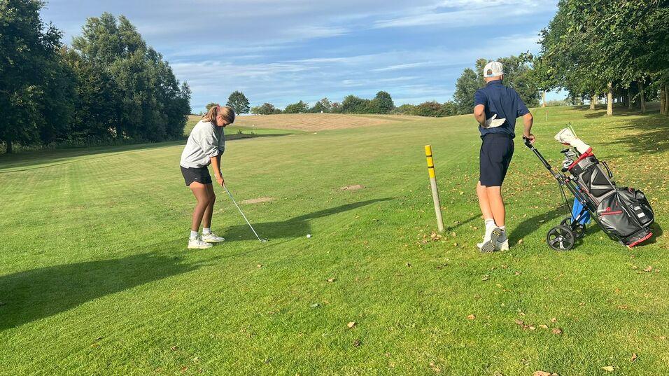 Zwei Golfer auf einer Rasenfl&auml;che, einer beim Schlag, der andere mit Golftasche im Hintergrund.