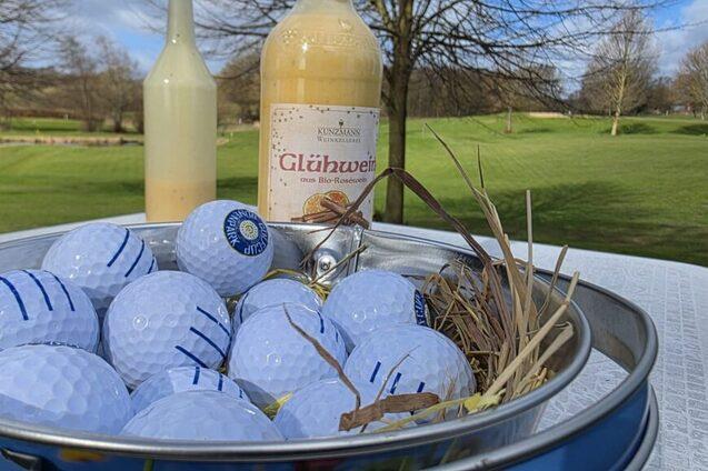 Ein Tisch voller blauer Golfbälle und einer Flasche Glühwein, im Hintergrund ein grünes Golfgelände und Bäume.