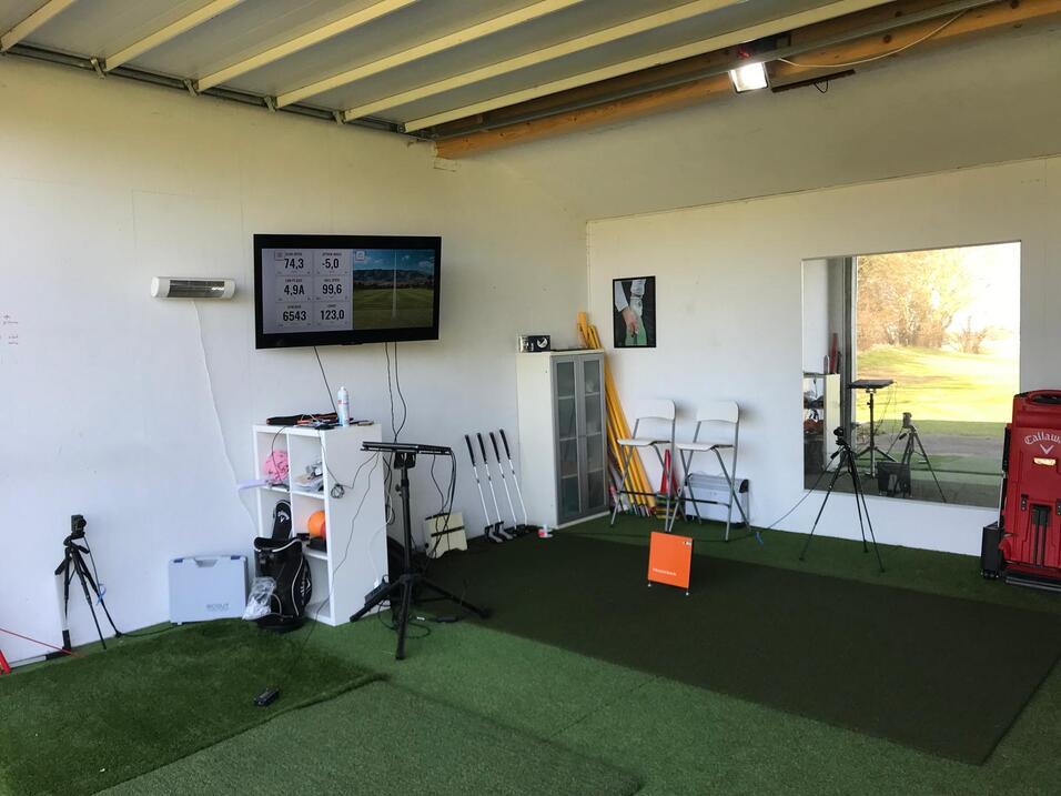 Innenansicht eines Golftrainingsraums mit Bildschirm, Putting-Grün und Übungsgeräten.