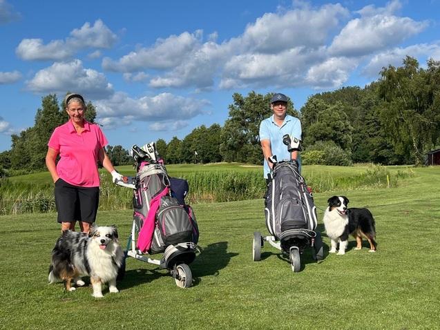 Zwei Frauen mit Golftaschen und zwei Hunden stehen auf einer Golfwiese unter blauen Himmel mit Wolken.