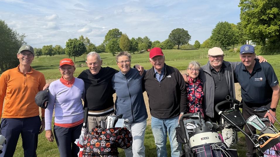 Gruppe von acht Golfern steht lächelnd auf einer Wiese mit Golftaschen neben sich und einem blauen Himmel im Hintergrund.